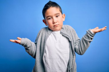 Küçük çocuk mavi izole edilmiş arka plan üzerine spor kazak giyiyor ve elleri ve kolları havada kafası karışmış bir ifade takınıyor. Şüphe kavramı.