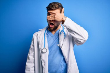 Sakallı, mavi gözlü, beyaz ceketli ve steteskoplu genç sarışın doktor yüzünü ve gözlerini kapayarak utangaç bir ifadeyle parmaklarının arasından bakıyor..