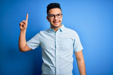 Yaz gömleği ve gözlük takan genç yakışıklı adam izole edilmiş mavi arka plan üzerinde başarılı bir fikirle parmağı işaret ediyor. Heyecanlı ve mutlu. Bir numara..