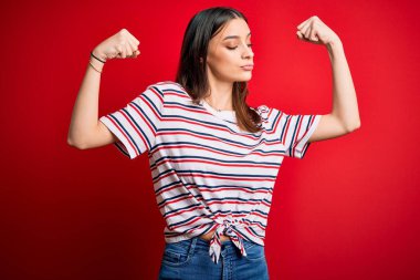 Genç, güzel, esmer, sıradan çizgili tişört giyen, kırmızı arka planda duran ve kollarında gururla gülümseyen bir kadın. Fitness konsepti.