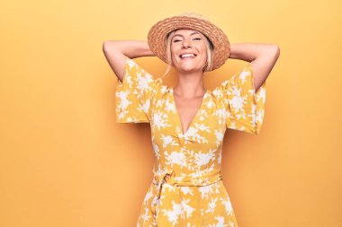 Sarışın, güzel bir kadın tatilde. Sarı arka planda giyinmiş. Kolları ve elleri başının arkasında, mutlu bir şekilde gülümsüyor.