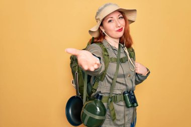 Sırt çantasıyla gezen, sarı arka planda şapkası olan genç, kızıl saçlı kadın mutlu bir şekilde avuç içi yardım ve kabullenme teklif ediyor..