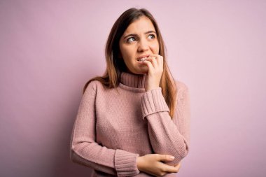 Pembe izole edilmiş arka plan üzerine balıkçı yaka kazak giyen güzel genç bir kadın elleri ağız yiyen tırnaklarla stresli ve gergin görünüyor. Endişe sorunu.