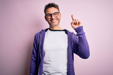Young handsome man wearing purple sweatshirt and glasses standing over pink background showing and pointing up with finger number one while smiling confident and happy. clipart