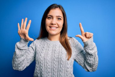 Güzel, genç bir kadın, mavi izole edilmiş arka planda dikilirken yün süveter giyer ve yedinci parmaklarıyla işaret ederken kendinden emin ve mutlu bir şekilde gülümser..