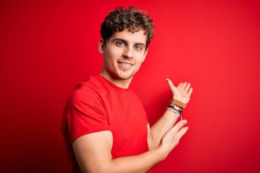 Kıvırcık saçlı, kırmızı arka planda sıradan tişörtler giyen genç sarışın adam gülümseyerek içeri girmeye davet ediyor.