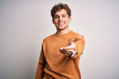 Sarışın, kıvırcık saçlı, beyaz arka planda sıradan bir süveter giyen neşeli bir avuç içi yardım eli uzatan ve kabul gören genç bir adam..