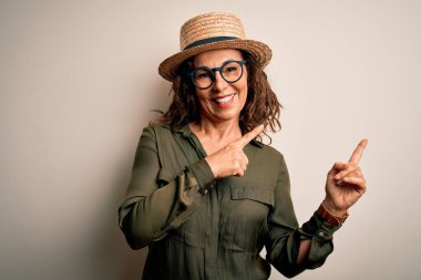 Orta yaşlı esmer kadın gözlüklü ve şapkalı. İzole edilmiş beyaz arka planda duruyor. Gülümsüyor ve kameraya bakıyor..