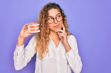 Güzel, mavi gözlü, mor arka planda plastik dişleri olan sarışın bir kadın. Ciddi bir yüz ifadesi var. Soru sormayı düşünüyor.