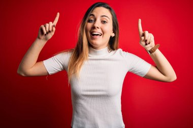 Mavi gözlü, güzel, beyaz tişörtlü, kırmızı arka planda gülümseyen, şaşkın, parmaklarıyla işaret eden ve kollarını kaldıran sarışın bir kadın..