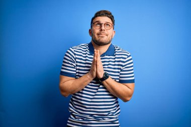 Mavi gözlü, gözlüklü ve mavi arka planda sıradan çizgili tişörtlü genç adam elleri bağlı yalvarıyor ve dua ediyor. Yüzünde umut ifadesi var. Çok duygusal ve endişeli. Yalvarıyorum..