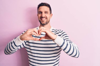Sıradan çizgili süveter giyen yakışıklı genç adam izole edilmiş pembe arka planda duruyor. Aşkla gülümsüyor, elleriyle kalp sembolü yapıyor. Romantik konsept.