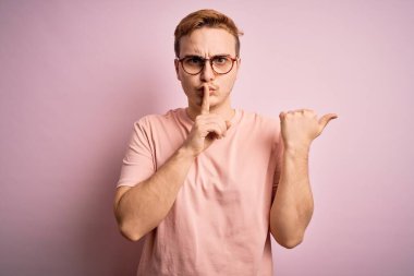 Genç, kızıl saçlı, günlük tişört giyen, pembe arka planda duran, sessiz olup dudaklarını yan tarafı işaret eden genç bir adam. Sessizlik ve gizli konsept.