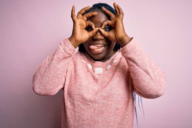 Afro-amerikalı örgü örgülü kadın pembe arka planda sıradan bir süveter giyiyor. Dürbün gibi jest yapıyor, gözleri parmaklarından bakıyor. Çılgın ifade.