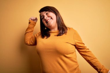 Güzel esmer ve cüsseli bir kadın izole edilmiş sarı arka planda günlük süveter giyiyor mutlu ve neşeli dans ediyor, hareketli ve kendinden emin bir şekilde müzik dinliyor.