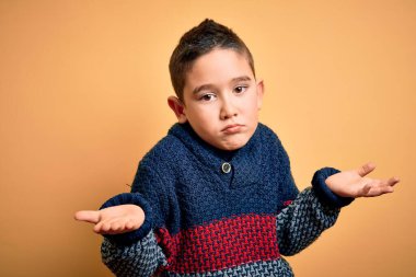 Sarı arka plan üzerinde kışlık süveter giyen küçük bir çocuk. Kolları ve elleri havada, kafası karışmış bir yüz ifadesi. Şüphe kavramı.