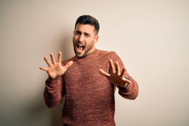 İzole edilmiş beyaz arka plan üzerinde duran sıradan bir süveter giyen genç ve yakışıklı bir adam korku ifadesi karşısında korkmuş ve dehşete düşmüş. El hareketlerini durdur, şok içinde bağır. Panik konsepti.