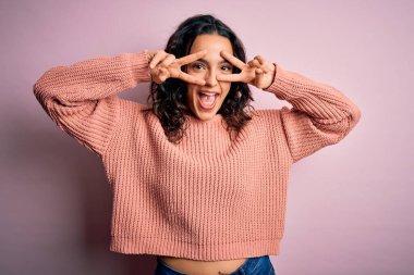 Kıvırcık saçlı, günlük süveter giyen, pembe arka planda barış sembolü çizen, parmaklarının üzerinde gülümseyerek zaferi gösteren genç ve güzel bir kadın.