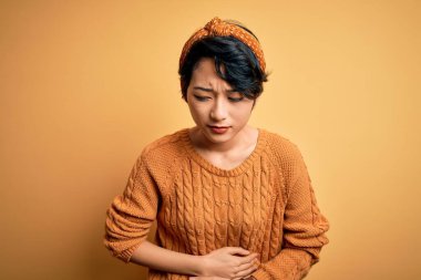 Genç ve güzel Asyalı kız gündelik süveter ve diyabet giyerek sarı arka planda duruyor. Midenin üstünde el üstünde duruyor. Çünkü hazımsızlık, acı veren hastalıklar iyi hissetmiyor. Ache kavramı.