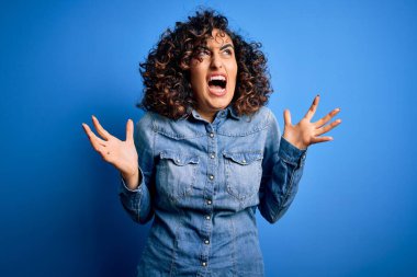 Genç ve güzel kıvırcık Arap kadın, sıradan bir kot gömlek giyiyor mavi arka planda deli deli deli bağırıyor ve agresif bir ifade ve kollarla bağırıyor. Hayal kırıklığı konsepti.