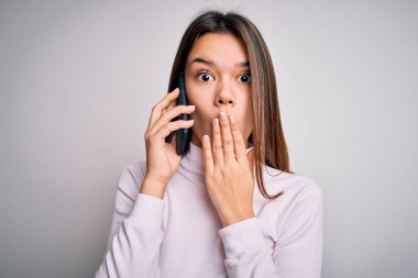 Güzel esmer kız, beyaz arka plan üzerinden akıllı telefondan konuşuyor. Ağzı kapalı, utancından ağzı açık, korkunun ifadesi, sessizlikten korkan, gizli bir konsept.