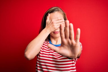 Genç ve güzel sarışın kadın, izole edilmiş kırmızı arka plan üzerine sıradan çizgili tişört giyip gözleri ellerle kaplıyor ve üzgün ve korku ifadeleriyle jest yapıyor. Utanç verici ve olumsuz bir kavram..