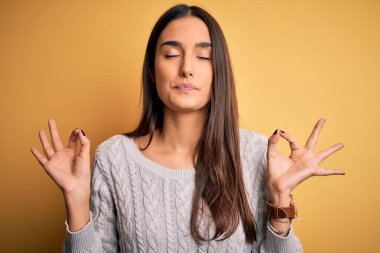 Sarı arka plan üzerine beyaz süveter giyen genç, güzel esmer kadın rahatla ve parmaklarıyla meditasyon hareketi yaparken gözleri kapalı gülümse. Yoga konsepti.