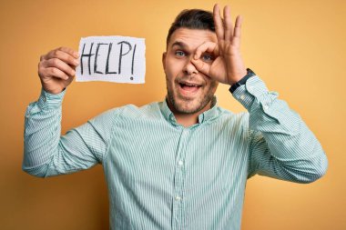 Mavi gözlü genç iş adamı sarı arka planda kağıt tutarak yardım istiyor gülümseyen yüzüyle tamam işareti yapıyor parmaklarının arasından bakıyor