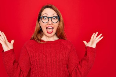 Genç, güzel, kızıl saçlı bir kadın kırmızı arka plan üzerinde gözlük ve süveter takarak çılgın ve başarıyı kollarını kaldırıp heyecanla haykırarak kutluyor. Kazanan konsept