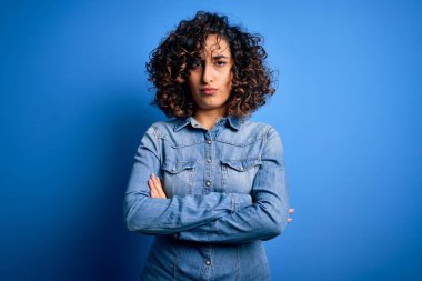 Genç ve güzel kıvırcık Arap kadın mavi arka plan üzerinde sıradan bir kot gömlek giyiyor şüpheci ve gergin, yüzünde çapraz kollu tasvip etmeyen bir ifade var. Negatif kişi.