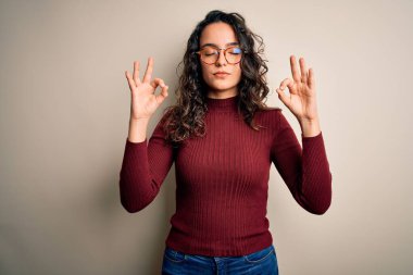 Kıvırcık saçlı, günlük süveter giyen, beyaz arka planda gözlük takan ve gözleri kapalı gülümseyen güzel bir kadın parmaklarıyla meditasyon hareketi yapıyor. Yoga konsepti.