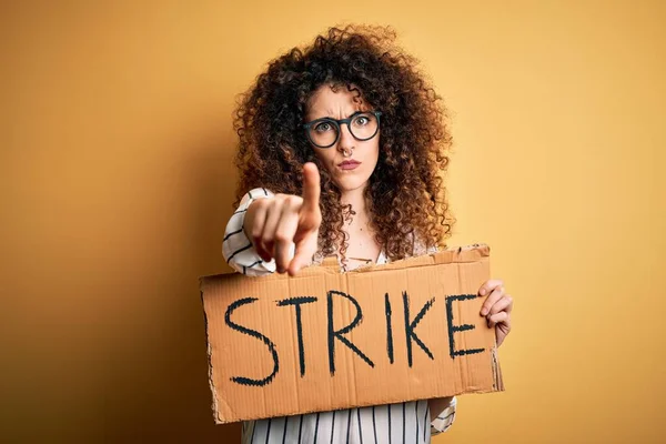 Young beautiful activist woman with curly hair and piercing protesting in strike holding poster pointing with finger to the camera and to you, hand sign, positive and confident gesture from the front