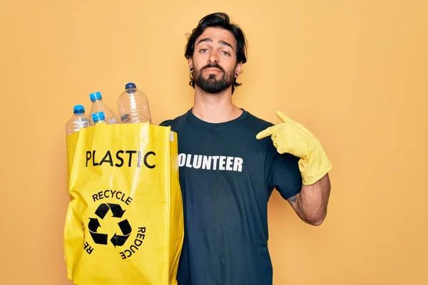 ボランティアのTシャツを着たハンサムなパニックボランティアの男性は 顔が指を指して驚くほどのペットボトルで袋を持っています — ストック写真