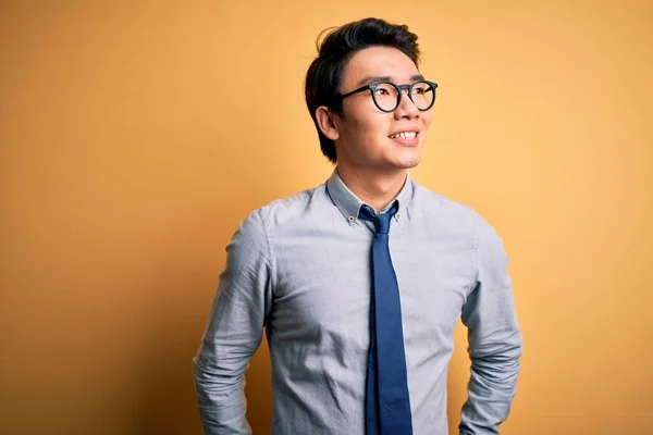 Young handsome chinese businessman wearing glasses and tie over yellow background looking away to side with smile on face, natural expression. Laughing confident.