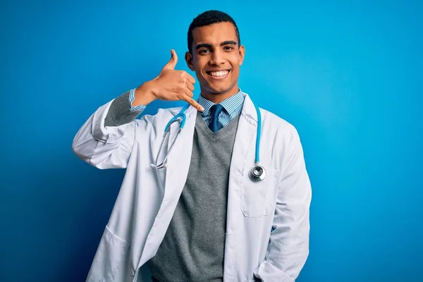 英俊的非洲裔美国医生 身穿外套 带着听诊器 带着蓝色背景的笑容 用手和手指做着电话手势 就像在电话里说话一样 交流概念 — 图库照片