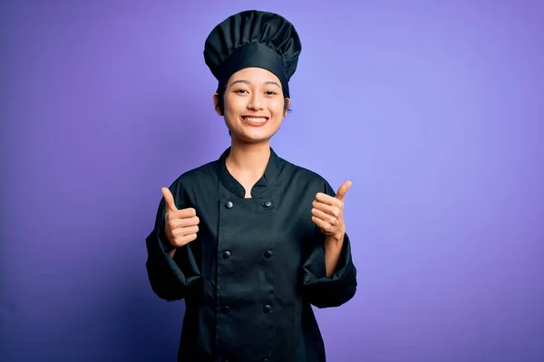 若い美しい中国のシェフの女性は 手で肯定的なジェスチャーを行う紫色の背景の成功のサインの上に炊飯器の制服と帽子を身に着けています 親指アップ笑顔と幸せ 陽気な表情と勝者のジェスチャー — ストック写真