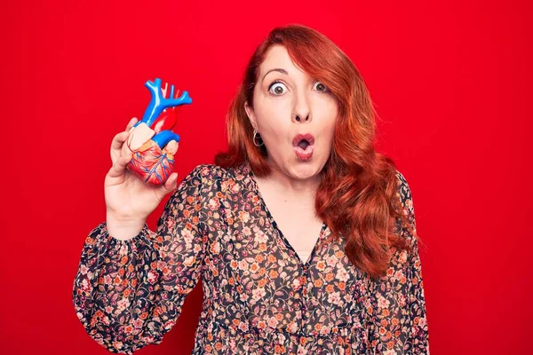 Young beautiful redhead woman asking for health care holding heart over read background scared and amazed with open mouth for surprise, disbelief face