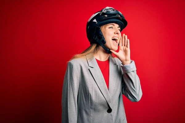 若い美しいブロンドのオートバイの女性は 赤い背景の叫びの上にオートバイのヘルメットを着用し 口の上に手で横に大声で叫んでいます コミュニケーションの概念 — ストック写真