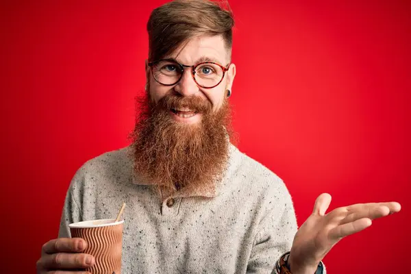 赤い背景の上に紙コップの上にテイクアウトコーヒーを飲むひげを持つアイルランドの赤毛の男は非常に幸せと興奮 大きな笑顔で叫んで勝利を祝う勝者式と手を上げ — ストック写真