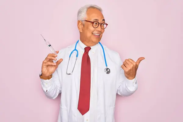 中年シニア白髪医者男保持薬針注射上のピンクの背景ポインティングと見せます上に親指で側に幸せな顔笑顔 — ストック写真