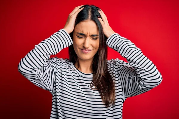 Genç, güzel esmer kadın kırmızı arka plan üzerine sıradan çizgili tişört giyiyor. Ağrı ve migren yüzünden baş ağrısı çekiyor. Ellerini başının üstüne koy..