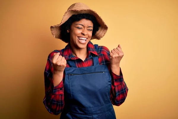若いアフリカ系アメリカ人のアフロ農家の女性は 巻き毛のエプロンを身に着けており 腕を上げて成功に興奮し 目を閉じて勝利笑顔を祝う帽子 勝者のコンセプト — ストック写真