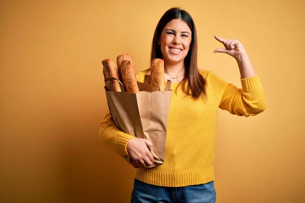 年轻美丽的女人拿着一袋黄色背景的健康的新鲜面包 微笑着 自信地做手势 手拿着小尺寸的标志 手拿着手指 手拿着相机 计量概念 — 图库照片