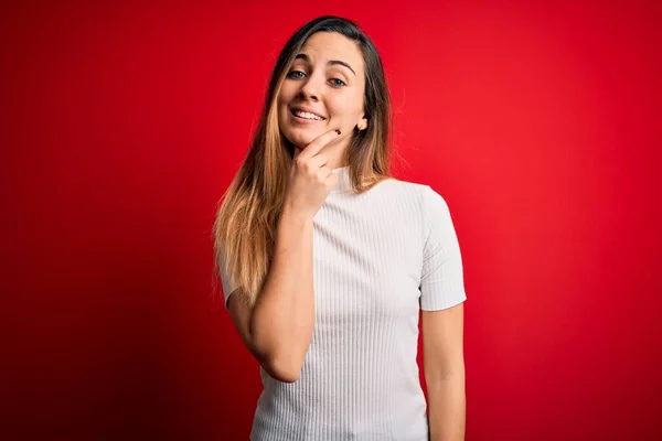 Beautiful blonde woman with blue eyes wearing casual white t-shirt over red background looking confident at the camera smiling with crossed arms and hand raised on chin. Thinking positive.
