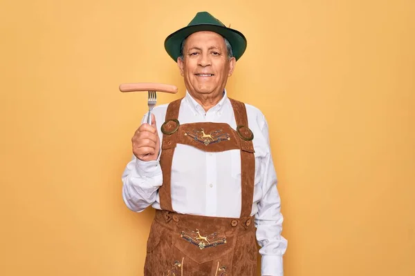 Senior grey-haired man wearing german traditional octoberfest suit holding fork with sausage with a happy face standing and smiling with a confident smile showing teeth