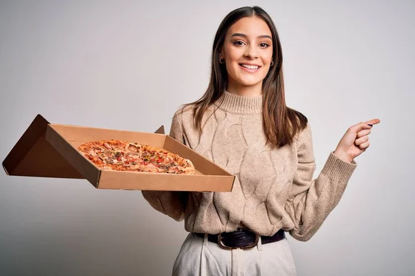 年轻美丽的黑发女人拿着装有意大利披萨的盒子 背景是白色的 她非常高兴地把手和手指指向旁边 — 图库照片