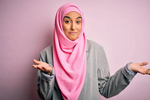 Young beautiful brunette businesswoman wearing pink muslim hijab and business jacket clueless and confused expression with arms and hands raised. Doubt concept.