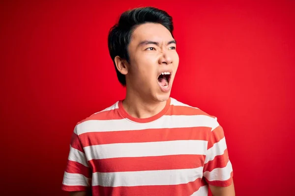 若いハンサムな中国人男性がカジュアルなストライプのTシャツを着て 赤の背景に立って怒っていると怒りを叫び 怒りで叫んで怒っている 激怒と積極的な概念 — ストック写真