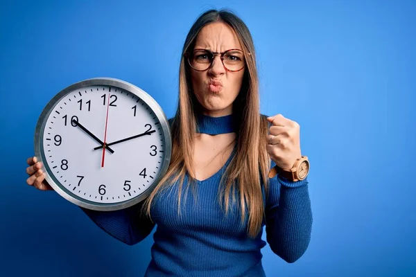 Young blonde woman with blue eyes holding big minute clock over isolated background annoyed and frustrated shouting with anger, crazy and yelling with raised hand, anger concept