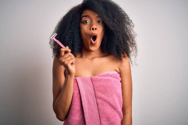 Young african american woman with afro hair in a towel holding female razor for body depilation scared in shock with a surprise face, afraid and excited with fear expression